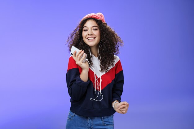 Tiro de cintura de encantadora y tierna mujer de pelo rizado en gorro y sudadera escuchando música mientras ...