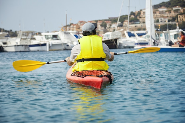 Tiro colorido de joven en canoa en un chaleco de natación amarillo con remo se detuvo en el aire. Hermoso paisaje de puerto encantador con yates blancos y casas de la ciudad cercana en la montaña.