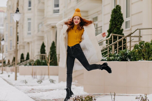 Tiro completo de linda modelo femenina en sombrero amarillo saltando en la calle Retrato al aire libre de mujer alegre jugando en invierno