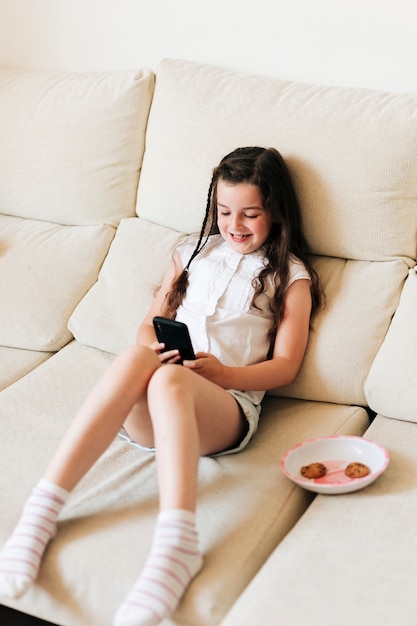 Foto gratuita tiro completo sonriente niña sentada y mirando el teléfono