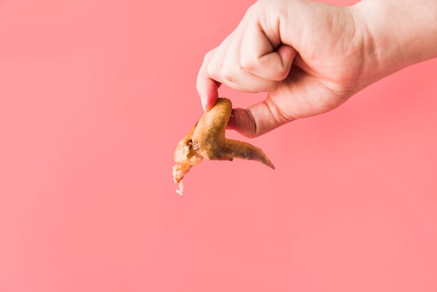 Tiro del detalle de la mano de la persona que sostiene las alas de pollo asadas delante del fondo rosado