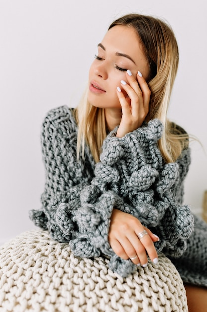 Tiro de estudio interior de hermosa mujer sensual con cabello rubio y ojos cerrados tocando su rostro con suéter de punto posando en la habitación blanca