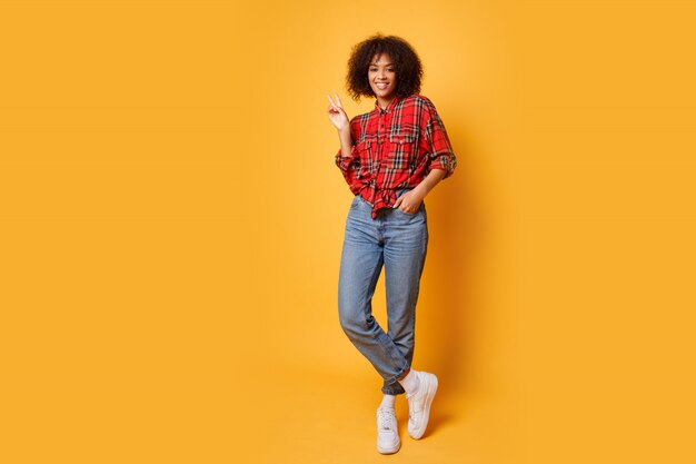 Tiro del estudio de la muchacha negra que salta con la expresión feliz de la cara en fondo anaranjado brillante. Usa jeans, zapatillas blancas y camisa roja.