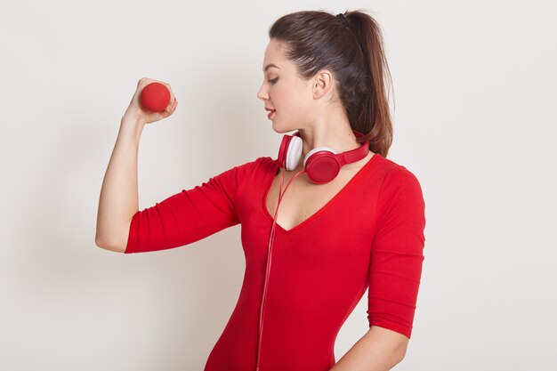 Tiro del estudio de la mujer caucásica sana con pesas que se resuelve aislado en blanco. Señora atractiva que muestra sus músculos, mirando en su brazo, Fitness, gimnasio, concepto de cuidado saludable.