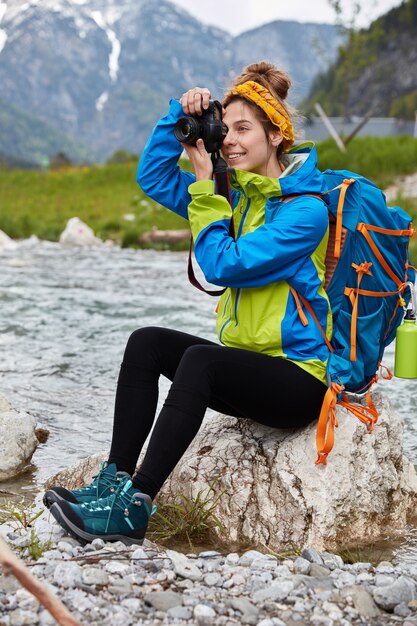 Tiro exterior vertical de mujer alegre hace fotos profesionales, se sienta en las rocas cerca del río de la montaña