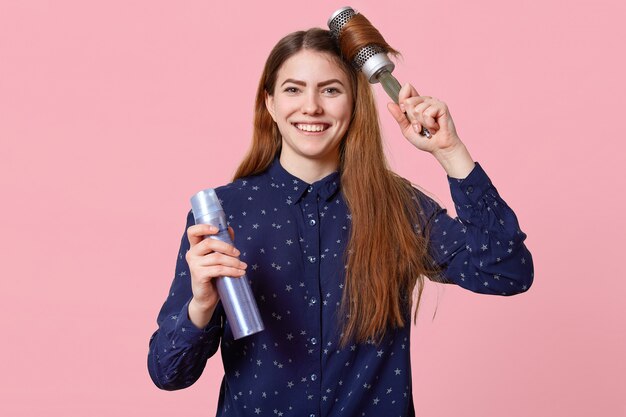 Tiro horizontal de apuesto sonriente mujer europea con cabello largo, utiliza cepillo para el cabello y laca para el cabello, vestido con elegante camisa