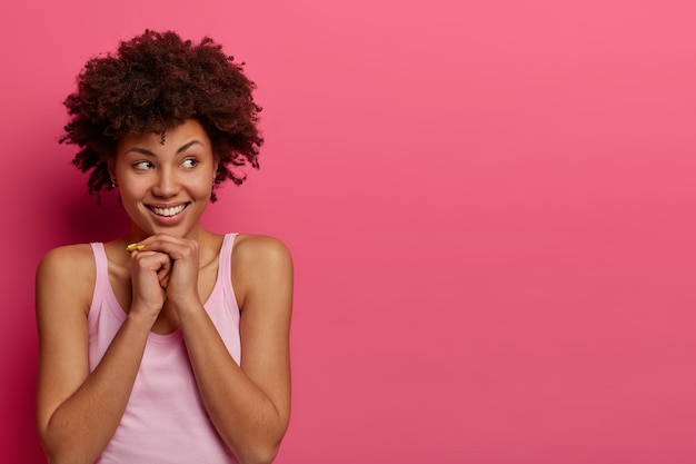 Tiro horizontal de una atractiva chica de piel oscura que mira a un lado con una sonrisa, mantiene las manos debajo de la barbilla, ve algo atractivo a la derecha, posa contra la pared rosa, espacio en blanco para su promoción