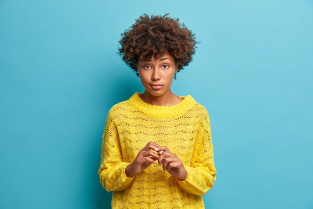 Tiro horizontal de atractiva mujer seria con cabello rizado mantiene las manos juntas y mira con curiosidad a la cámara escucha atentamente al interlocutor vestido con un jersey casual aislado en la pared azul