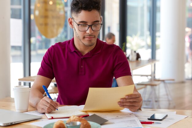 Tiro horizontal de banquero serio sostiene papel, escribe ideas creativas para el desarrollo de un negocio bancario exitoso, sostiene bolígrafo para escribir en el bloc de notas, rodeado de modernos aparatos en la cafetería