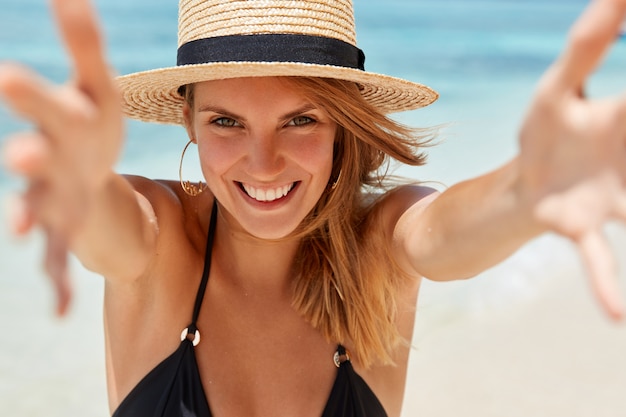 Tiro horizontal de bastante joven turista se divierte en la playa, extiende las manos como si fuera a abrazar a alguien, expresa felicidad, se olvida de todos los problemas en un lugar paradisíaco para un descanso maravilloso