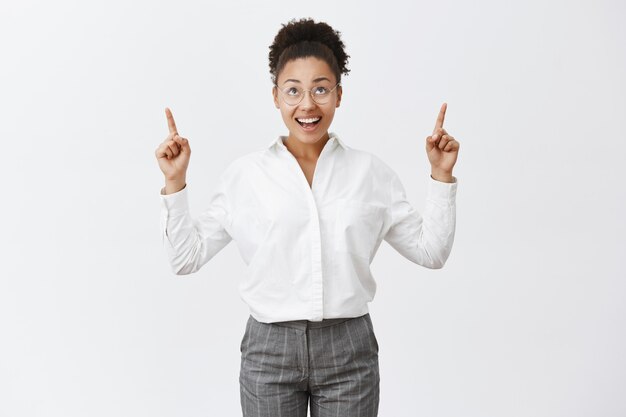 Tiro horizontal de encantadora mujer africana despreocupada con cabello rizado en gafas, apuntando hacia arriba con los dedos índices, mirando al cielo con expresión cálida y feliz, teniendo un gran día en la oficina con compañeros de trabajo