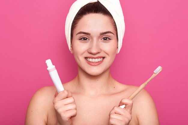 El tiro horizontal de la encantadora mujer caucásica sonriente aislada en la pared rosada del estudio, lista para cepillarse los dientes, sosteniendo la pasta de dientes y el cepillo de dientes en las manos, tiene una piel suave perfecta. Concepto de higiene.
