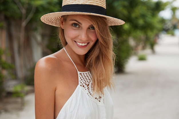 Tiro horizontal de feliz hermosa mujer con sonrisa positiva, viste sombrero de verano y vestido blanco, camina al aire libre, disfruta de un buen descanso durante las vacaciones y el clima cálido y soleado. Vacaciones y estilo de vida