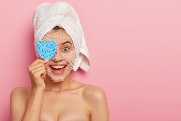 Tiro horizontal de feliz modelo de mujer joven cubre los ojos con una esponja cosmética, limpia la cara con una mascarilla exfoliante de sal de mar blanca natural, visita el salón de spa, tiene una piel sana y suave, cuerpo desnudo, modelos de interior