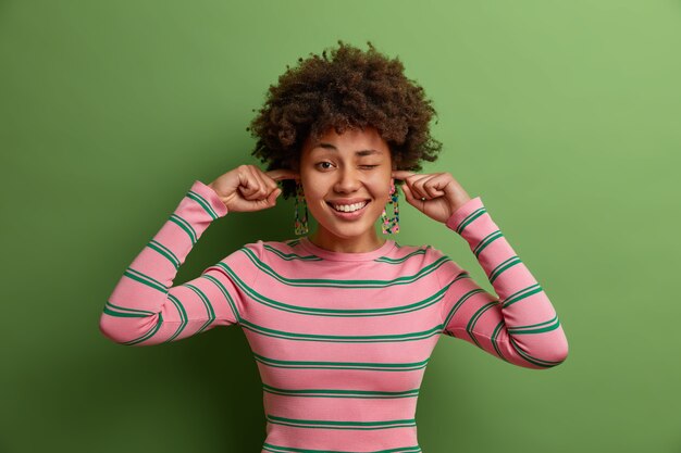 Tiro horizontal de feliz mujer étnica tapona los oídos con los dedos, escucha música alta en la fiesta, guiña los ojos y posa en el interior, vestida con un jersey de rayas casual. La mujer ignora el ruido proveniente de los vecinos