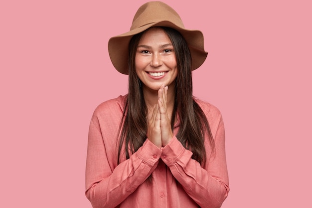 Tiro horizontal de hermosa mujer sonriente con expresión satisfecha, mantiene las manos juntas de alegría