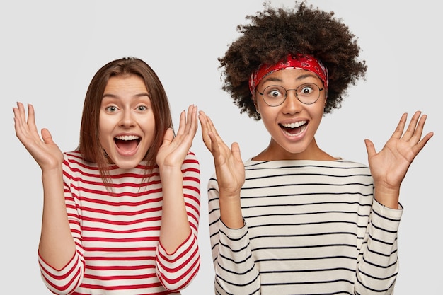 Foto gratuita tiro horizontal de hermosas hembras asombradas se dan la mano con felicidad, se reúnen con su mejor amigo, vestido informalmente, aislado sobre una pared blanca. concepto de personas, raza y emociones positivas.