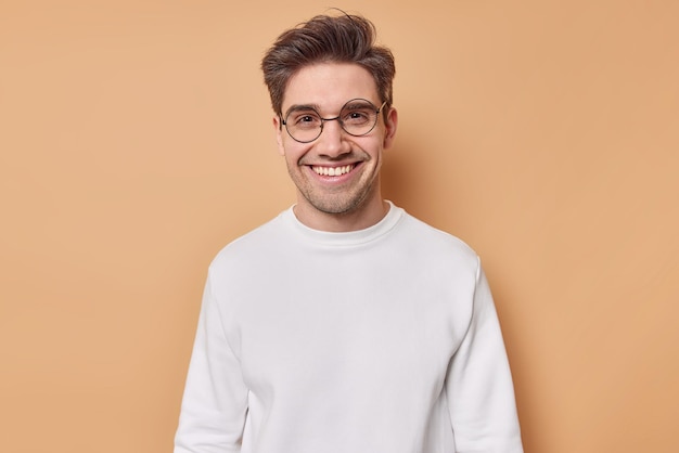 Tiro horizontal de hombre adulto alegre guapo contento de conocer a su mejor amigo tiene una conversación agradable con el interlocutor usa gafas redondas y un jersey blanco casual aislado sobre fondo beige