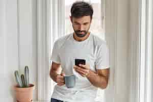 Foto gratuita tiro horizontal de hombre guapo con barba utiliza moderno dispositivo de teléfono inteligente, bebidas calientes, posa cerca del alféizar de la ventana interior.