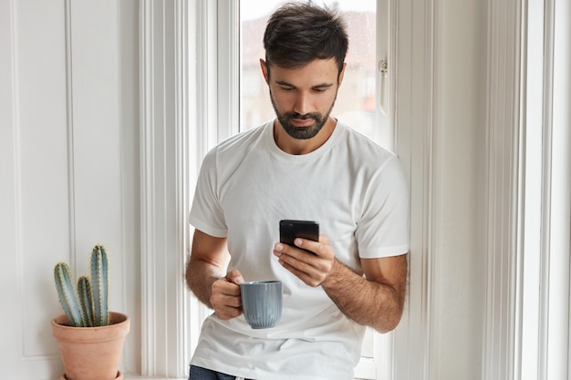 Tiro horizontal de hombre guapo con barba utiliza moderno dispositivo de teléfono inteligente, bebidas calientes, posa cerca del alféizar de la ventana interior.