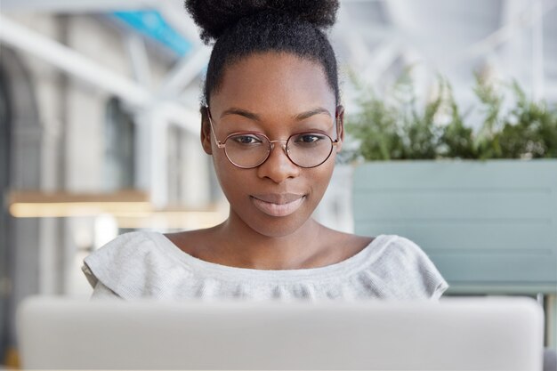 Tiro horizontal de mujer afroamericana enfocada en gafas redondas lee información en la computadora portátil, navega por internet.