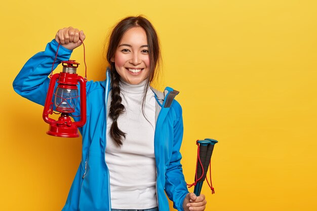 Tiro horizontal de mujer asiática alegre con trenza oscura, sostiene lámpara de gas y bastones de trekking, vestida con ropa deportiva, tiene una sonrisa positiva sobre la pared amarilla.