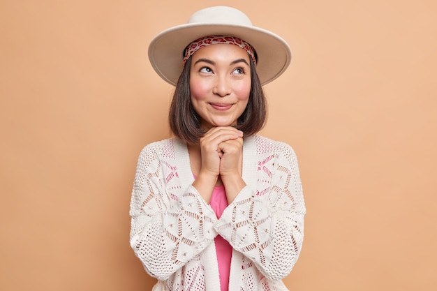 Tiro horizontal de mujer asiática soñadora mantiene las manos debajo de la barbilla piensa en algo agradable viste fedora jersey de punto blanco hace planes en mente aislado sobre pared beige