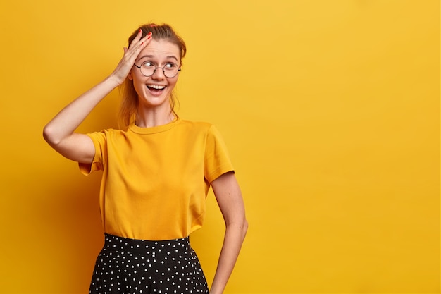 Tiro horizontal de mujer bastante alegre mira hacia otro lado mantiene la mano en la cabeza sonríe ampliamente usa camiseta básica y falda anteojos ópticos aislados sobre pared amarilla espacio en blanco para su anuncio