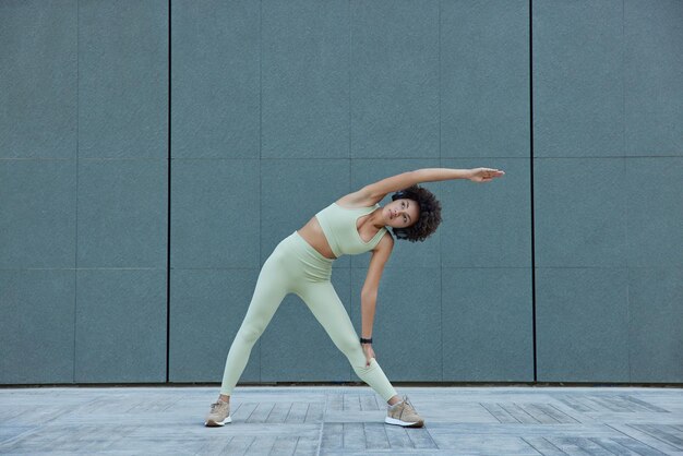 Tiro horizontal de mujer delgada deportiva en ropa activa se inclina a un lado ejercicios contra la pared gris en blanco al aire libre se mantiene saludable mantiene buena forma física tiene entrenamiento regular sigue el régimen matutino diario