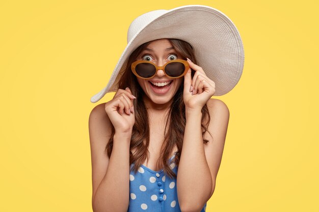 Tiro horizontal de mujer muy atractiva con cabello oscuro, mira con sorpresa y felicidad, mantiene la mano en el borde de las sombras, modelos en traje de verano sobre pared amarilla. Concepto de belleza y emociones