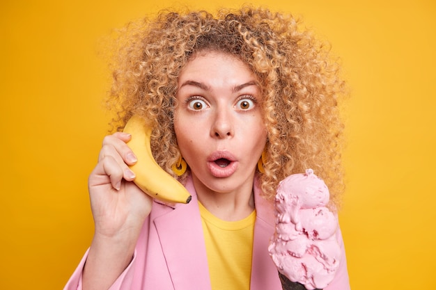 Tiro horizontal de mujer rizada sorprendida que mira fijamente los ojos saltones contiene la respiración mantiene el plátano como si el teléfono cerca de la oreja come un helado delicioso durante el día caluroso recibe demasiadas calorías tiene una nutrición poco saludable