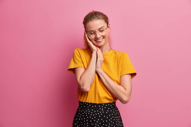 Tiro horizontal de niña bonita sonriente mantiene las manos cerca de la cara cierra los ojos con placer