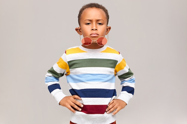 Tiro horizontal de niño afroamericano guapo fresco con expresión facial segura manteniendo las manos en la cintura, elegantes tonos rosados redondos cayendo por su nariz. Concepto de infancia y moda