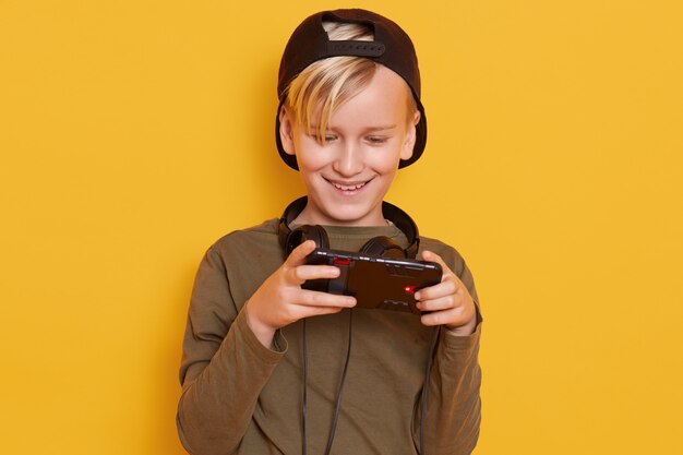 Tiro horizontal del niño pequeño con gorra negra y sudadera verde, posando con el teléfono celular en las manos, niño de moda jugando juegos en línea.
