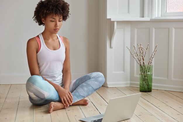 Tiro horizontal de relajada mujer de piel oscura sentada en posición de loto, mira videos de entrenamiento, aprende yoga, vestida con ropa casual, posa en un piso de madera en una habitación vacía. Concepto de tecnología y descanso