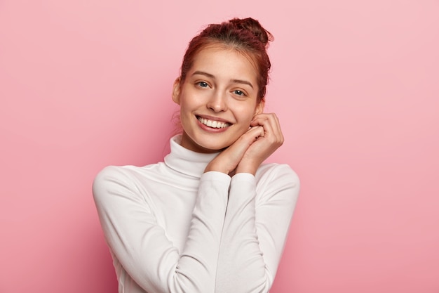 Tiro horizontal de tierna sonrisa inclina la cabeza, toca la piel limpia y sana, mira con adoración, tiene una sonrisa con dientes, viste coqueteos de cuello alto blanco y posa en interiores en una pared rosada