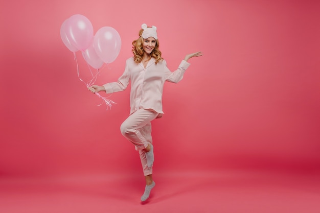 Tiro integral de cumpleañera en calcetines grises posando. Retrato de joven sonriente en pijama de seda saltando con globos de color rosa.