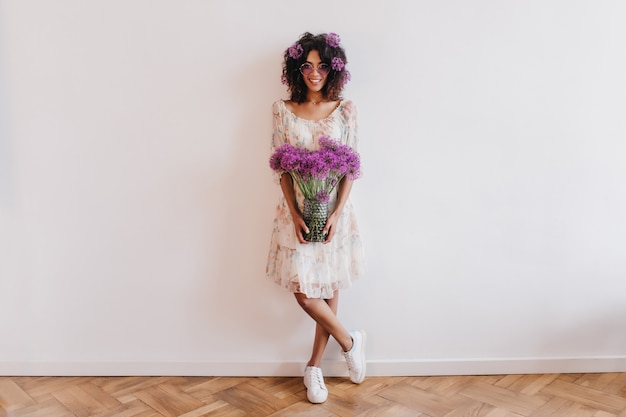 Foto gratuita tiro integral de niña africana en zapatillas posando con jarrón de flores. retrato interior de dama negra delgada de pie con las piernas cruzadas y sonriendo.