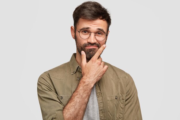 Tiro interior de desconcertado, vacilante, sin afeitar, chico sostiene la barbilla y duda, levanta las cejas, tiene una expresión desorientada, viste una camisa elegante, aislada sobre una pared blanca. Personas, emociones, concepto de estilo de vida