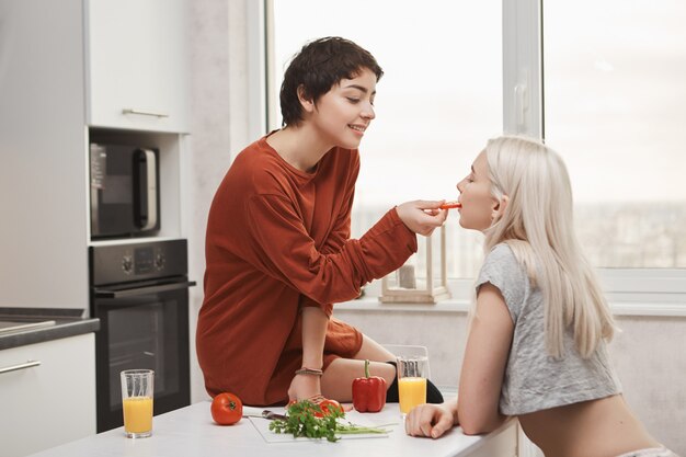 Tiro interior dulce y lindo de la mujer de pelo de camisa caliente que alimenta a su novia mientras está sentado en la mesa de la cocina y prepara el desayuno. Juego previo de la joven pareja sensual de chicas