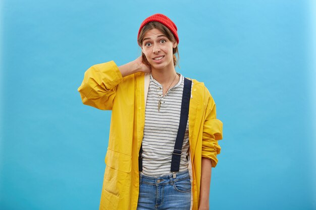 Tiro interior joven mujer con chaqueta suelta amarilla, overoles de mezclilla y sombrero rojo manteniendo su mano en el cuello para caminar en el bosque y recoger setas o bayas. Mujer vestida casualmente en la pared azul