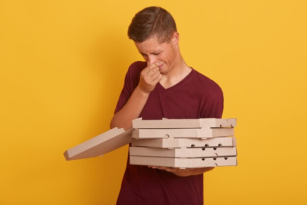 Tiro interior del joven repartidor guapo con mal olor malcriado pixxa, hombre con cajas de cartón, posando aislado en amarillo, chico con ropa casual. Concepto de entrega.