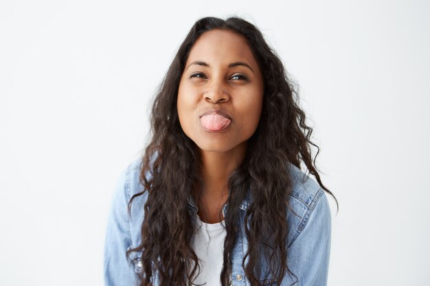 Tiro interior de mujer afroamericana bonita morena haciendo una mueca mostrando su lengua con expresión complacida. Mujer joven bastante juguetona con piel oscura posando en la pared blanca.