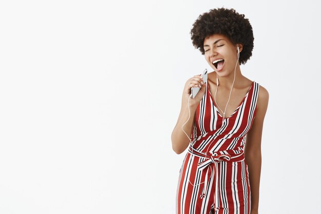 Tiro interior de mujer afroamericana despreocupada creativa y feliz con peinado afro sosteniendo teléfono inteligente como micrófono cantando música mientras escucha canciones en auriculares con alegría