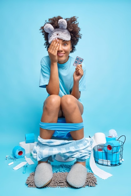 Foto gratuita tiro interior de mujer joven de pelo rizado complacida cubre el ojo con la mano sostiene poses de analgésicos en la taza del inodoro usa bragas tiradas hacia abajo en las piernas sufre de diarrea aislada sobre fondo azul.