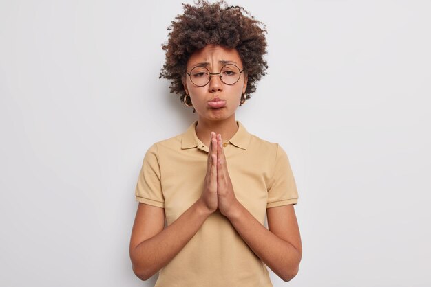 Tiro interior de mujer de pelo rizado disgustada mantiene las palmas presionadas juntas hace una solicitud con expresión triste, labios, lleva gafas redondas, camiseta beige informal aislada sobre fondo blanco