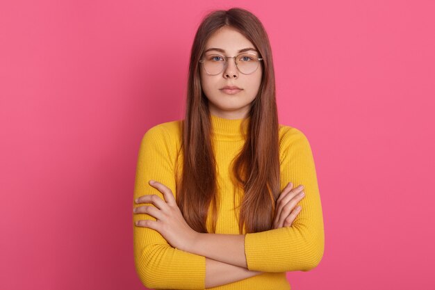 Tiro interior de pie femenino serio o enojado con los brazos cruzados. Chica joven con el pelo largo viste camisa amarilla