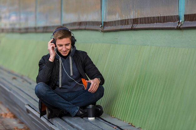 Tiro largo sonriente joven escuchando música con auriculares en un banco