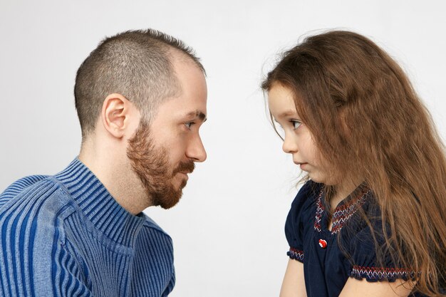 Tiro lateral de apuesto joven con rastrojo posando en la pared blanca con su pequeña hija, mirándose, teniendo alguna discusión