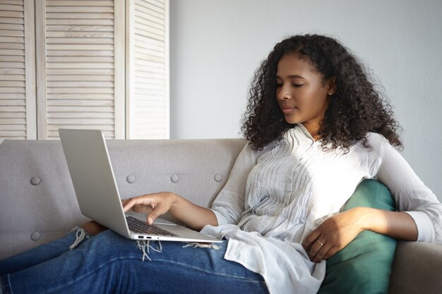 Tiro lateral de atractiva joven afroamericana viendo películas en línea en su computadora portátil genérica mientras se relaja en el sofá en casa solo después del trabajo. Personas, tecnología moderna y ocio